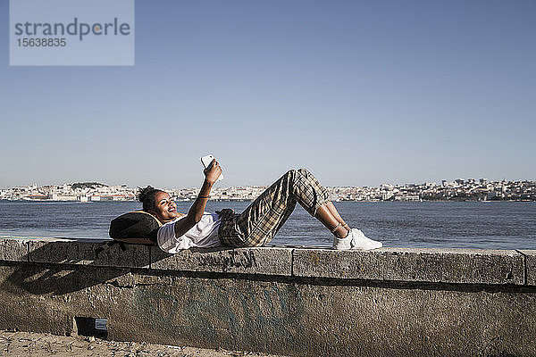 Junge Frau  die mit ihrem Handy an einer Wand am Wasser liegt  Lissabon  Portugal