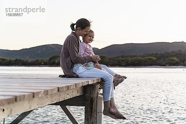 Glückliche Mutter mit Tochter sitzt bei Sonnenuntergang auf einem Steg