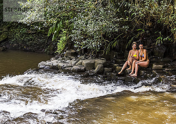 Zwei Frauen in Bikinis sitzen an einem der Twin Falls Wasserfälle und schauen in die Kamera; Haiku  Maui  Hawaii  Vereinigte Staaten von Amerika