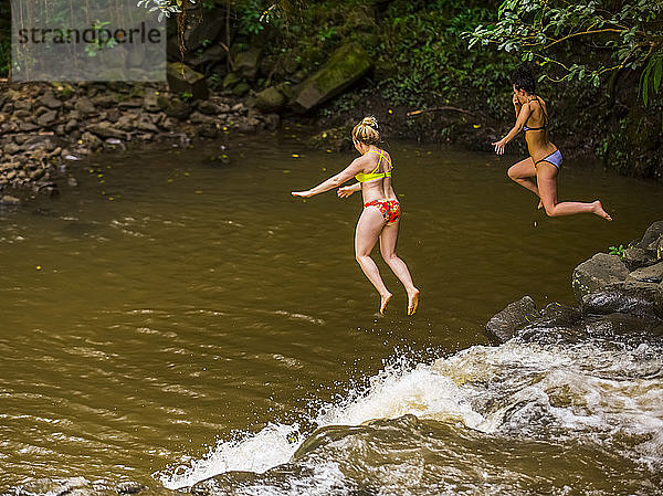 Zwei Frauen in Bikinis beim Klippenspringen von der Spitze eines der Twin Falls Wasserfälle; Haiku  Maui  Hawaii  Vereinigte Staaten von Amerika