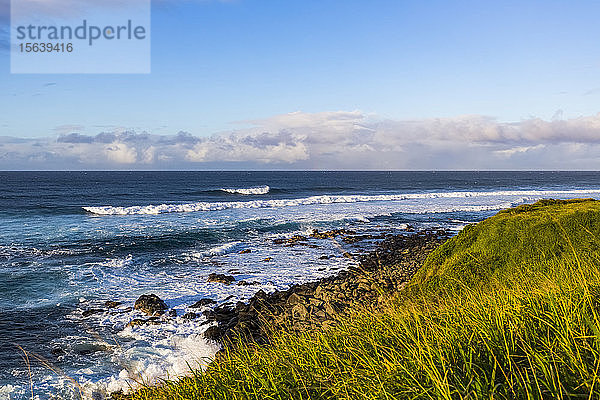 Meereswellen  die bei Sonnenuntergang an die Felsen und das grasbewachsene Ufer rollen  mit einer ruhigen Meereslandschaft und Wolken am Horizont des Ho'okipa Lookout bei Paia; Maui  Hawaii  Vereinigte Staaten von Amerika