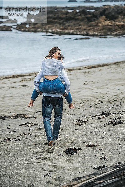 Ein junges Paar am Strand  die junge Frau wird auf dem Rücken des jungen Mannes getragen; Wellington  Nordinsel  Neuseeland