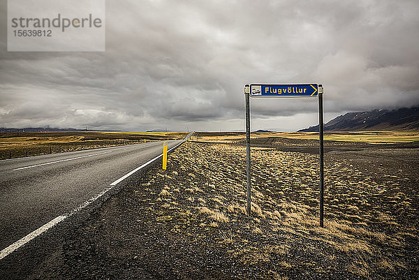 Straßen- und Tundra-Landschaft mit Flughafenschild am Straßenrand; Island