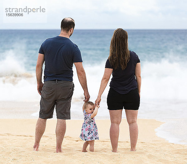 Mutter und Vater halten die Hände ihrer kleinen Tochter  während sie auf den Ozean hinausschauen; Maui  Hawaii  Vereinigte Staaten von Amerika