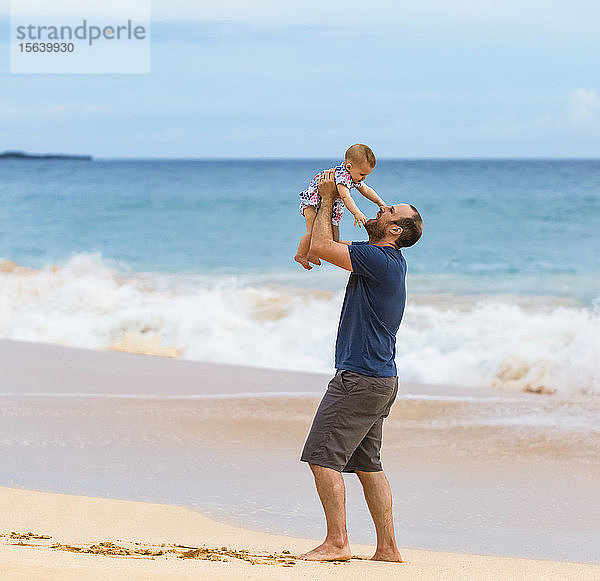 Vater hält seine kleine Tochter in die Luft  während er am Strand neben dem Ozean steht; Maui  Hawaii  Vereinigte Staaten von Amerika