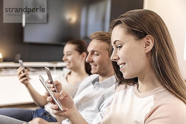 Freunde sitzen auf der Couch  lächelnd  schauen auf ihr Smartphone  Deutschland  Europa