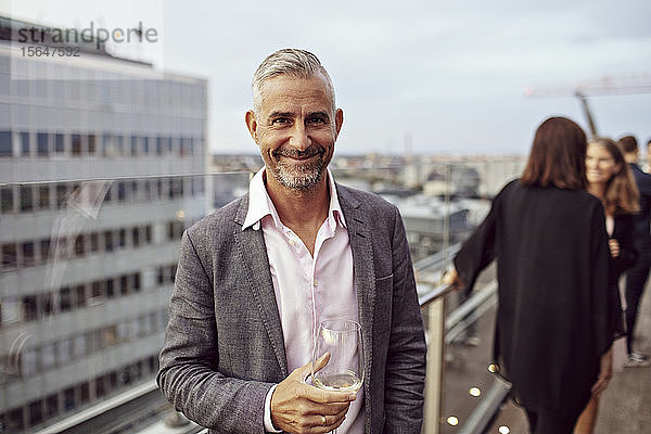 Porträt eines lächelnden Geschäftsmannes mit Weinglas in der Hand und Mitarbeitern im Hintergrund auf der Terrasse