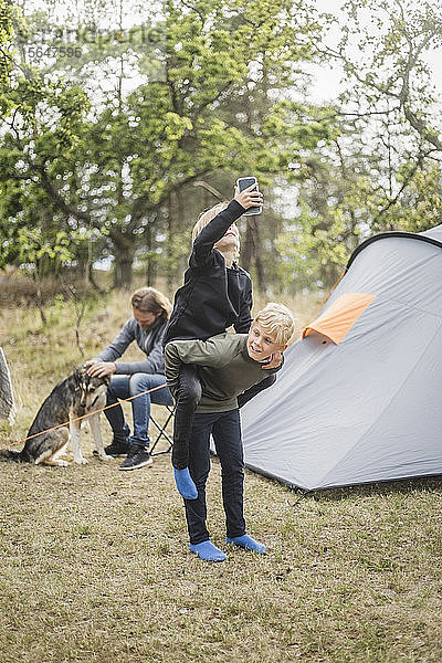 Junge nimmt Schwester mit Smartphone huckepack  während Mann beim Zeltcamping mit Hund spielt