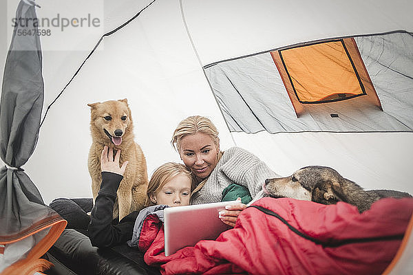 Mutter und Tochter beobachten digitales Tablet  während sie mit Haustieren im Zelt auf dem Campingplatz liegen