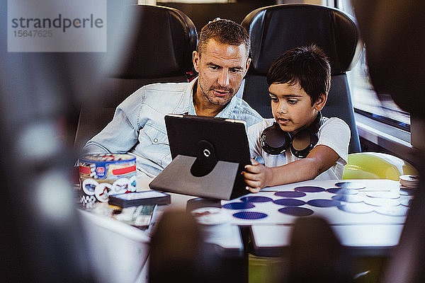 Mittelgroßer erwachsener Mann mit Sohn betrachtet digitales Tablet  während er im Personenzug sitzt