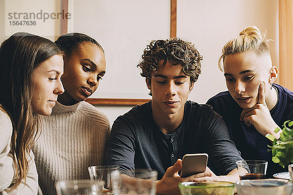 Teenager zeigt Freunden sein Handy  während er im Zimmer am Tisch sitzt
