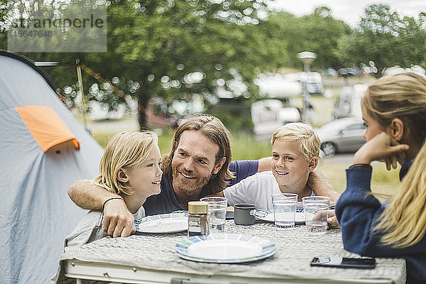 Lächelnder Vater sitzt mit Kindern am Tisch auf einem Campingplatz