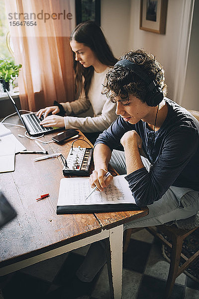 Hochwinkelansicht eines Teenagers  der mit Kopfhörern Musik hört  während ein Freund einen Laptop am Tisch im Haus benutzt