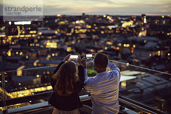 Rückansicht von männlichen und weiblichen Berufstätigen  die die Stadt bei Sonnenuntergang von der Terrasse aus mit dem Handy fotografieren