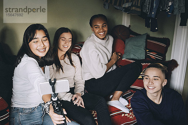 Lächelndes Teenager-Mädchen  das sich mit Freunden auf einem Einbeinstativ selbstständig macht  während es sich zu Hause vergnügt