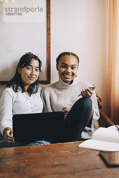 Porträt von lächelnden Teenager-Mädchen mit Laptop und Smartphone  die zu Hause auf dem Sofa sitzen