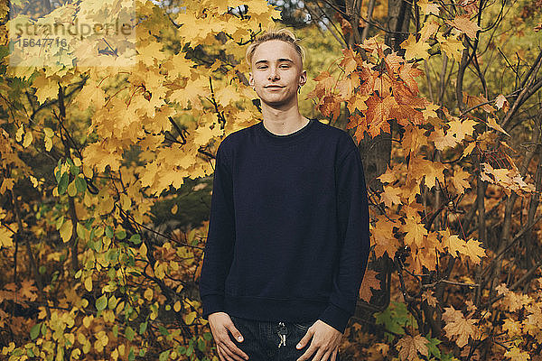 Zuversichtlicher Teenager mit blonden Haaren steht im Herbst vor Ahornbäumen