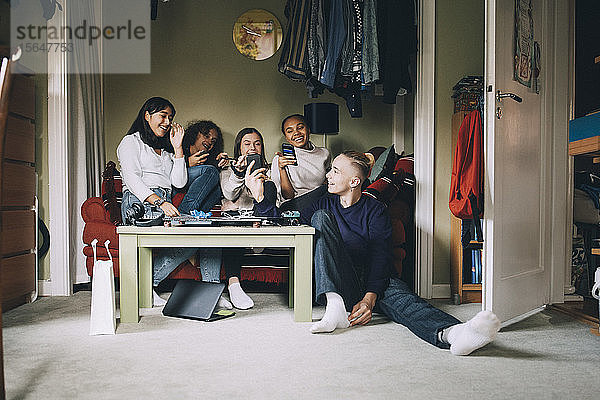 Fröhliche Freunde unterhalten sich beim Blick auf das Smartphone im Wohnzimmer