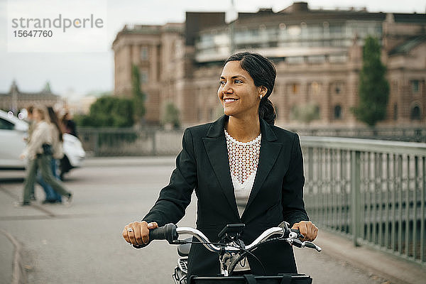 Lächelnde Geschäftsfrau mit Fahrrad auf einer Brücke in der Stadt