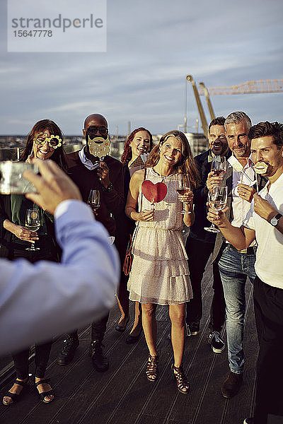 Geschäftsmann fotografiert glückliche Mitarbeiter mit Weingläsern und Requisiten auf Büroterrasse in Party