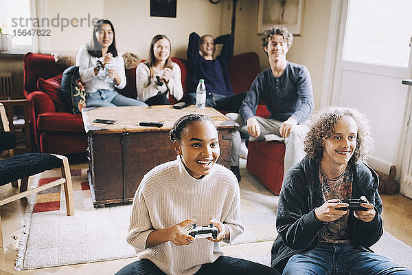Glückliche Teenager spielen Videospiele  während Freunde auf dem Sofa im Wohnzimmer sitzen
