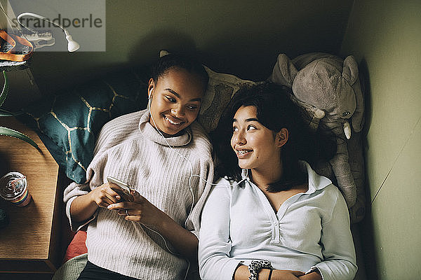 Direkt über der Aufnahme von lächelnden Teenager-Mädchen  die zu Hause auf dem Bett liegend Musik mit dem Handy hören
