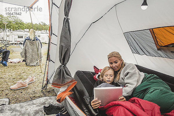 Mädchen sieht sich ein digitales Tablet an  während sie mit der Mutter im Zelt auf dem Campingplatz liegt