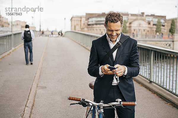 Lächelnder Geschäftsmann  der ein Mobiltelefon benutzt  während er mit dem Fahrrad auf einer Brücke in der Stadt steht