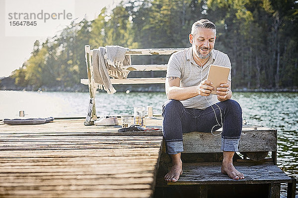 Lächelnder  reifer Mann benutzt digitales Tablett mit In-Ohr-Kopfhörern  während er auf dem Steg über dem See sitzt