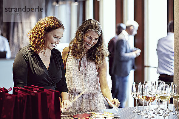Lächelnde Geschäftsfrauen schauen auf Requisiten durch Weingläser auf dem Tisch in der Büroparty