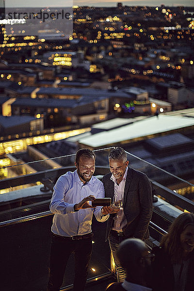 Schrägaufnahme von fröhlichen Geschäftsleuten  die sich mit einem Smartphone selbstständig machen  während sie nach der Arbeit auf der Terrasse stehen und beleuchtet sind