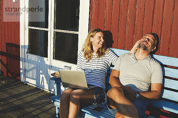 Glückliche Frau  die im Sommer mit einem Laptop spielt  während sie mit einem Mann auf einer Bank auf der Veranda spielt