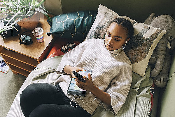 Hochwinkelansicht eines weiblichen Teenagers  der Musik hört  während er zu Hause am Bett ein Smartphone benutzt