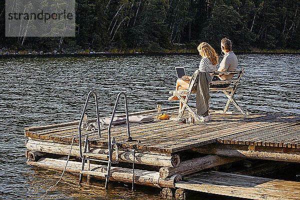 Mann und Frau mit Laptop  während sie im Sommer auf einer Bank über dem Pier am See sitzen