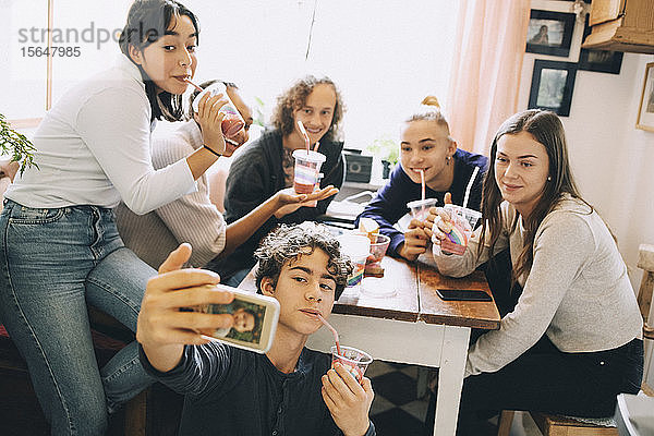 Teenager Junge nimmt Selfie mit Freunden über Handy mit  während er zu Hause einen Smoothie genießt