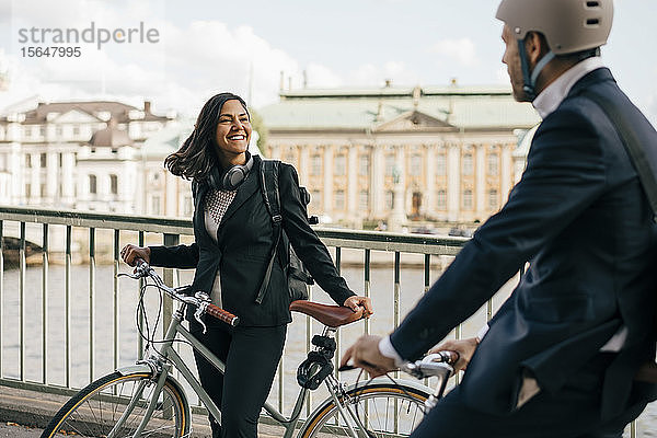 Lächelnde Geschäftsfrau mit Fahrrad schaut auf einen Kollegen  der auf der Brücke radelt