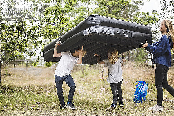 Teenager-Mädchen in voller Länge vergnügt sich mit Geschwistern  während sie zusammen eine aufblasbare Matratze auf dem Campingplatz tragen