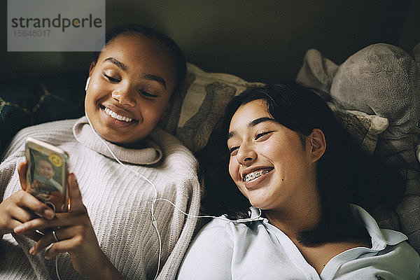 Glückliche weibliche Teenager  die zu Hause im Bett liegend auf einem Smartphone Musik hören