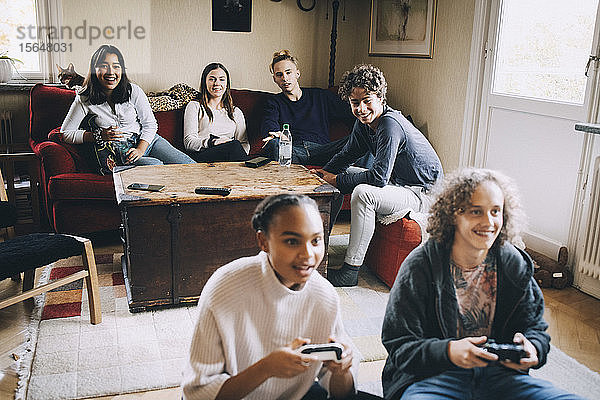 Lächelnde Männer und Frauen spielen Videospiele  während Freunde zu Hause im Hintergrund sitzen