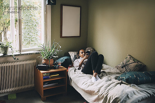 Teenager-Mädchen in voller Länge benutzt Smartphone  während sie zu Hause im Bett liegt