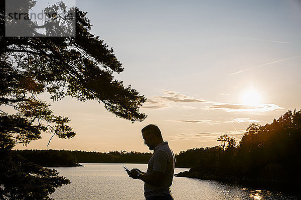 Scherenschnitt eines Mannes mit einem Smartphone  der bei Sonnenuntergang am See steht