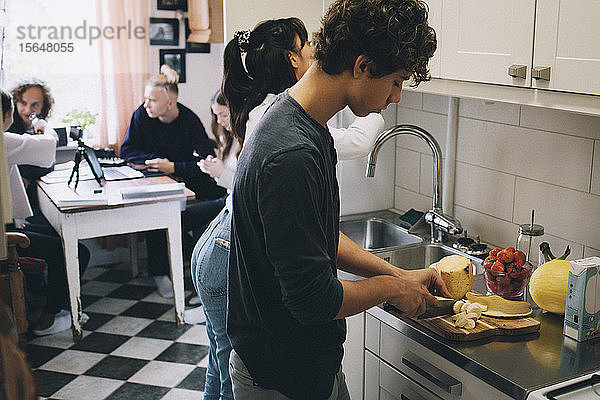 Teenager mit einer Frau  die Früchte an der Küchentheke schneidet  während Freunde im Hintergrund sitzen