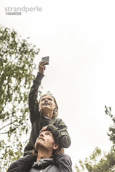 Niedrigwinkelansicht eines Mädchens  das durch ein Mobiltelefon fotografiert  während es auf den Schultern des Vaters gegen den Himmel sitzt