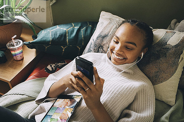Hochwinkelansicht eines lächelnden Teenager-Mädchens  das Musik hört  während es zu Hause am Bett ein Mobiltelefon benutzt
