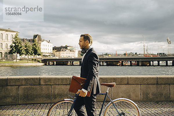 Seitenansicht eines Geschäftsmannes zu Fuß mit dem Fahrrad auf einem Fußweg in der Stadt gegen den Himmel