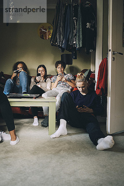 Männliche und weibliche Teenager in voller Länge  die zu Hause sitzen und Mobiltelefone benutzen