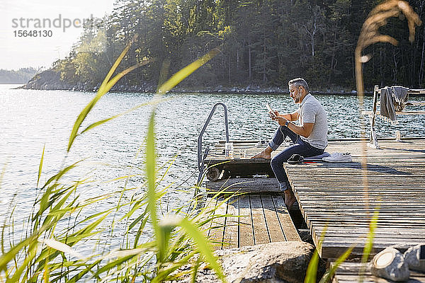 Mann in voller Länge mit digitalem Tablett auf Holzsteg über einem See im Wald sitzend