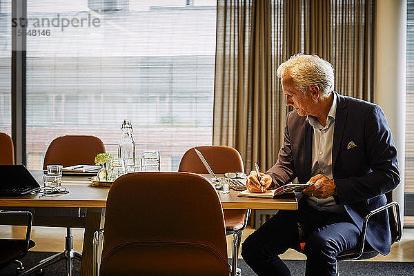 Reife Juristen schreiben im Tagebuch  während sie ihren Laptop am Konferenztisch im Sitzungssaal benutzen