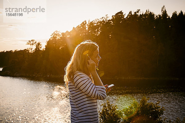 Seitenansicht einer mittleren erwachsenen Frau  die ein Mobiltelefon benutzt  während sie bei Sonnenuntergang am See steht