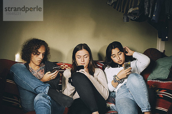 Abgelenkte männliche und weibliche Freunde benutzen Mobiltelefone  während sie zu Hause auf dem Sofa sitzen